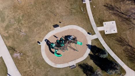 Drone-looking-down-over-a-playground-as-it-slowly-moves-up-to-reveal-people-are-at-play,-walking-and-enjoying-the-new-spring-sunny-day