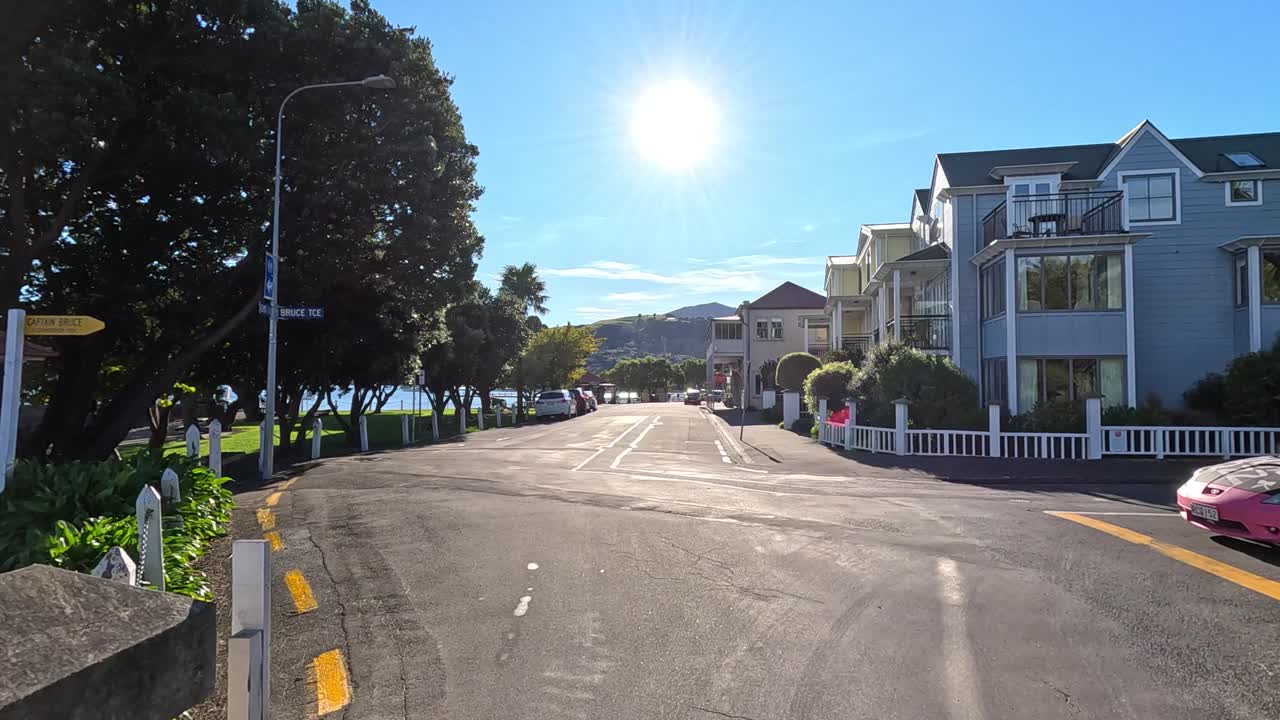 A serene drive along Akaroa's waterfront with vibrant sunlight and picturesque houses, captured in a smooth, continuous motion