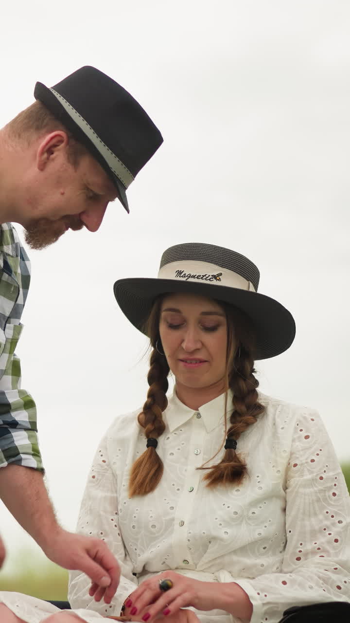 A craftsman in a checked shirt delicately adjusts the hand of a smiling woman wearing a white dress and hat, seated in a lush green field