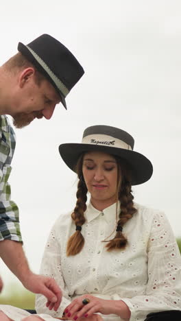 a craftsman in a checked shirt delicately adjusts the hand of a smiling woman wearing a white dress and hat, seated in a lush green field