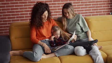 young women read a book together to a small preschool girl on sofa