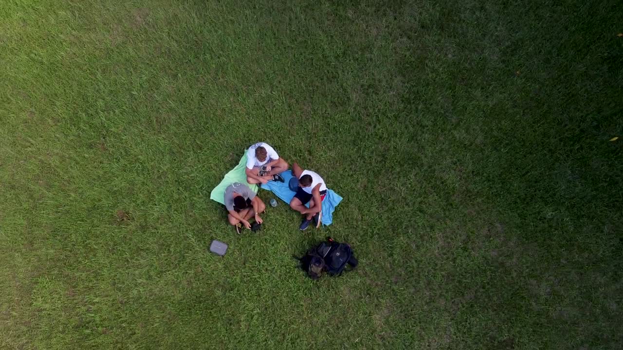 Ascending top down shot of three men sitting on grass field in park during sunny day - Iguazu, Argentina