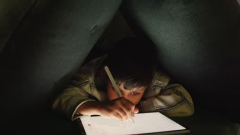 Young-Boy-In-Home-Made-Camp-Made-From-Cushions-Playing-And-Drawing-With-Digital-Tablet-And-Stylus-At-Night-1