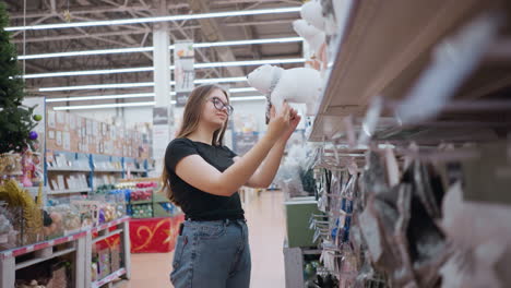 una joven con gafas y una camisa negra observa pensativamente un juguete blanco de peluche de oso polar en un estante ocupado de una tienda de vacaciones, rodeada de una variedad de decoraciones festivas y una brillante iluminación aérea