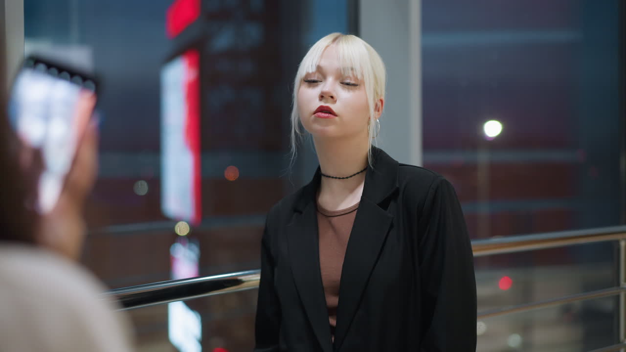 Elegant blonde lady standing indoors with eyes closed posing for photo near glass window as city lights reflect outside creating calm stylish atmosphere in modern shopping mall