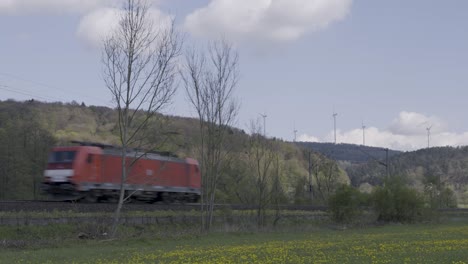 Freight-train-speeding-past-vibrant-yellow-wildflowers-and-bare-trees-under-a-cloudy-sky,-with-distant-wind-turbines-on-a-hill