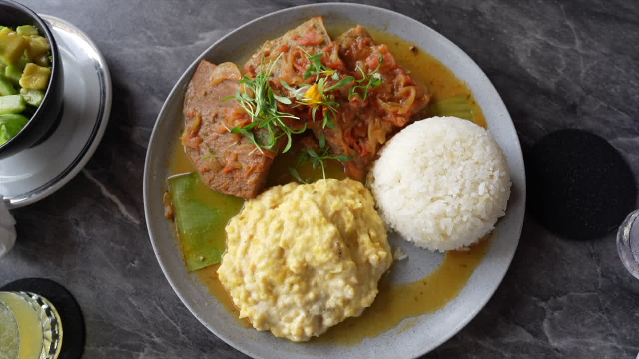 A delectable plate featuring Colombian sobrebarriga with plantain smash and rice, served on a gray surface. The vibrant dish is garnished, creating a colorful, inviting scene