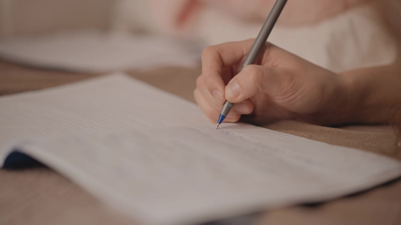 Close view of child hand holding ballpoint pen and writing on lined notebook while resting on bed, showing fresh ink trail, finger pressure, pen angle, soft light, calm bedroom routine
