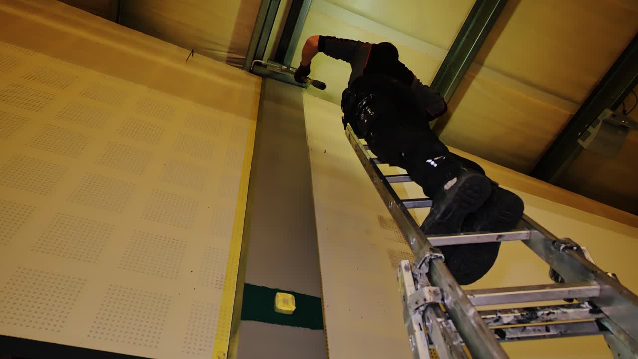 Low-lit interior scene as craftsman paints industrial post from steep height