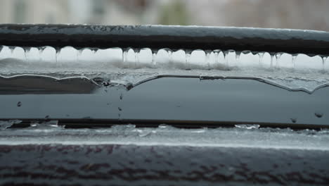 close-up of vehicle roof coated with a thick layer of ice and snow, featuring icicles forming along the edges of a black metal rail, with a blurred wintery urban background