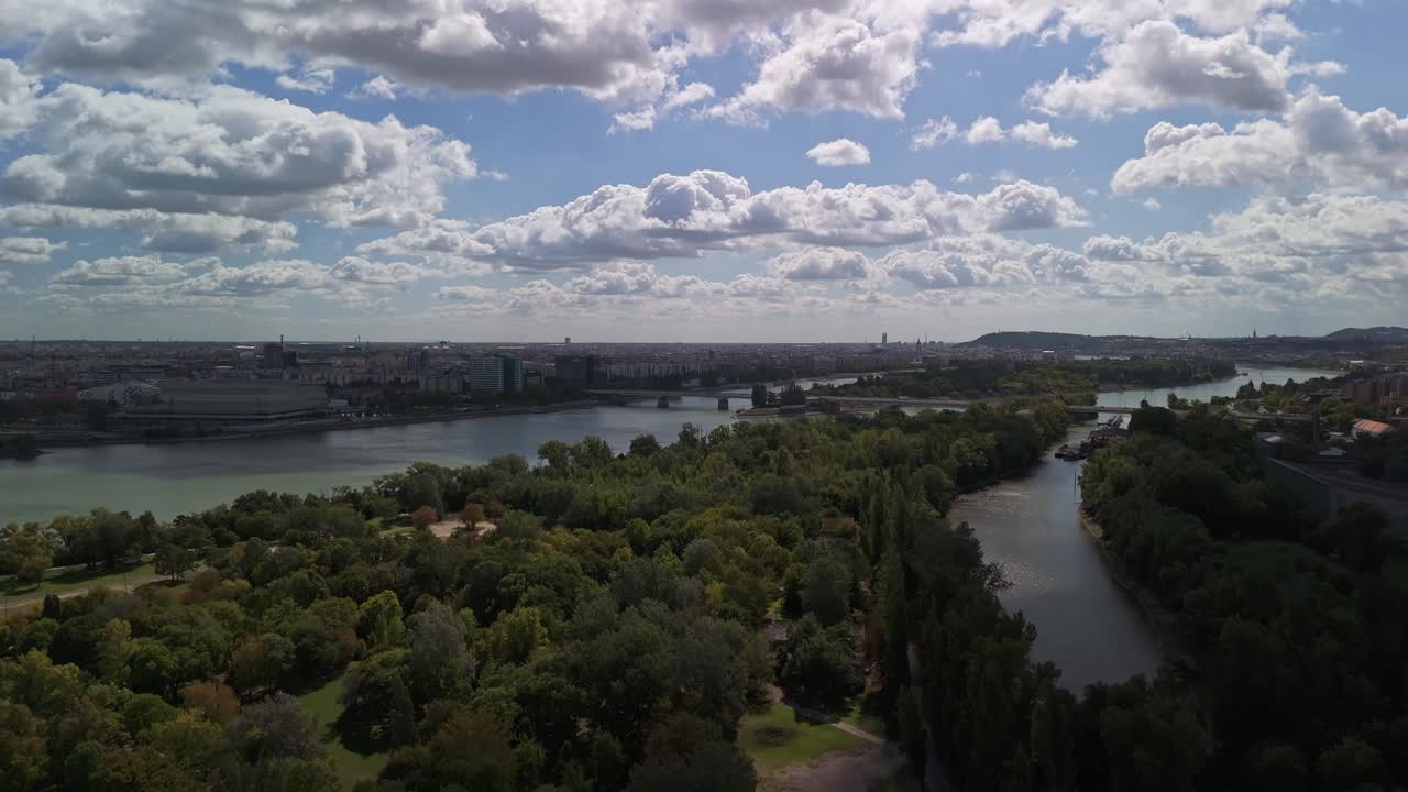 Vista aérea ascendente de Budapeste, Hungria, a partir de Óbudai-sziget durante o outono, mostrando a Ilha Margarida ao longo do Rio Danúbio, com paisagem urbana, pontes e vegetação sob um céu azul nublado