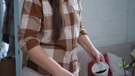 top down view of plant lover filling white plastic planter cup with dark soil using green scoop, focused facial expression, surrounded by potted plants and tools under soft natural light indoors