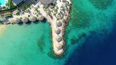 Tropical-resort-palm-frond-umbrellas-overlook-Caribbean-blue-ocean-water,-aerial-top-down