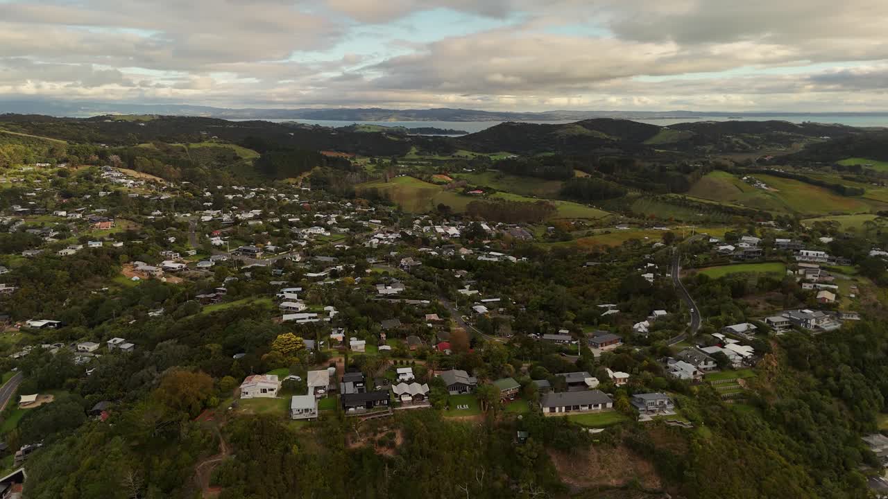 Luxury mansion on cliff edge of Coastline on Waiheke Island, new Zealand. Aerial wide shot. Green island with small town and homes. Mountain range in distance. Cloudy day on tropical island.