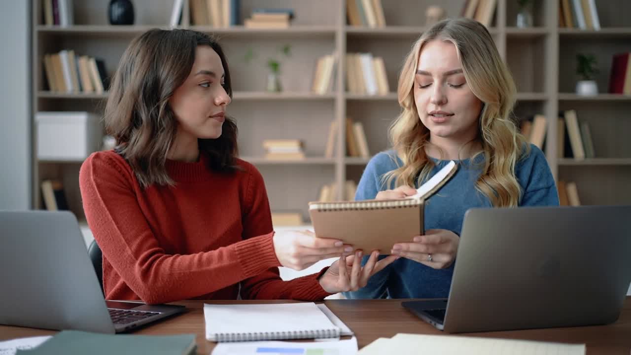 Video verticale: Donne in maglioni rossi e blu si protendono per prendere un blocco note e lo condividono alla scrivania, esaminando.