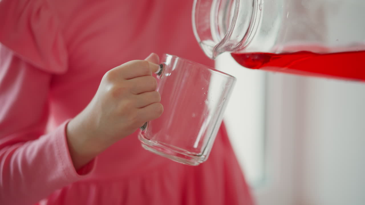 Hand view of little one sitting by sunny window handling glass jug to pour bright red drink into clear cup, sunny scene highlighting translucent liquid flowing smoothly as small hands grip handle