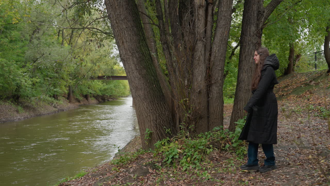 Žena stojící u řeky pod stromy, dlouhý kabát a boty na břehu, zamyšlená pauza u velkého kmene, zatažená obloha a zelená koruna, jemný proud vody a klidná nálada, osamělý portrét u řeky
