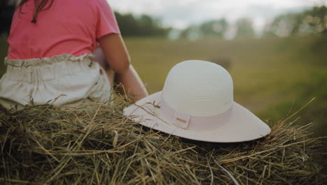 elegant white sun hat rests on hay bale while little girl in pink scratches her leg, holding a flower, warm sunlight bathes the countryside, with blurred trees in distance