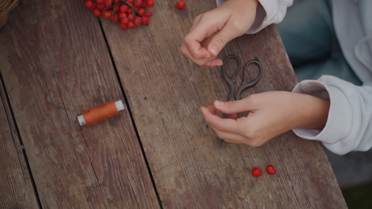 Hands threading red rowan berries garland, closeup of artisan creating seasonal decoration with spool of twine on rustic wooden table, cozy autumn mood, slow meditative movements, detailed sleeve