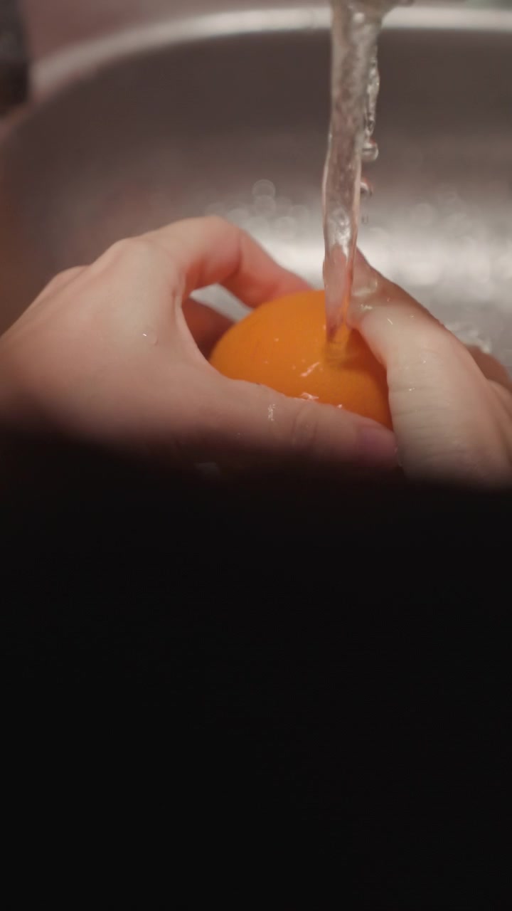 Close up of hands washing orange under running water in sink. Person rubs fruit to clean all sides with steady stream. Stainless steel kitchen setting with focus on hand movements