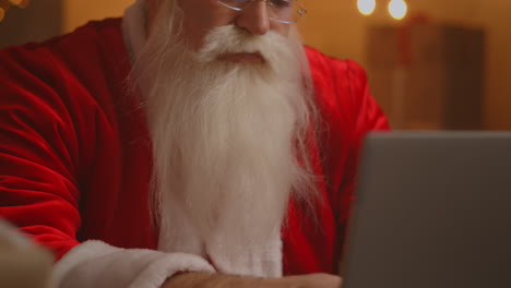 Real-Santa-Claus-using-new-technology-for-communication-with-children-receiving-mail-or-wish-list.-Cheerful-working-on-laptop-and-smiling-while-sitting-at-his-chair-with-fireplace-and-Christmas-Tree.-High-quality-4k-footage