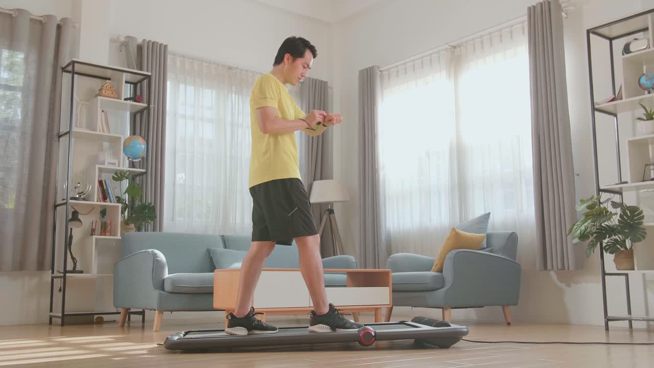 Side View Of Asian Man Training On Walking Treadmill And Looking Smart Watch At Home