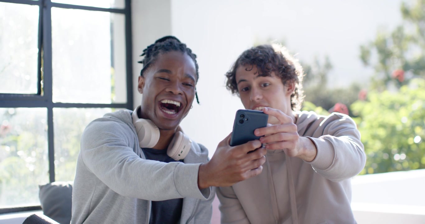 Diverse male friends on couch by window left man extending phone for selfie checking pics laughing
