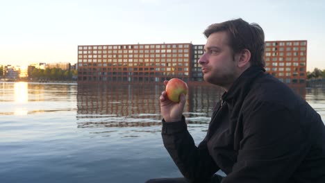 young handsome man in a black jacket eating a red apple on an urban pier in amsterdam during sunset