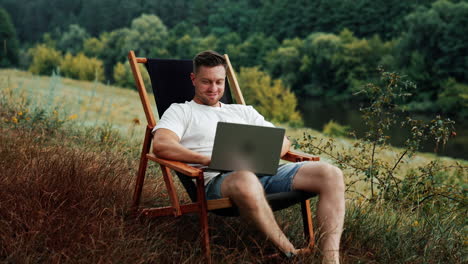 happy male freelancer working in the beautiful nature. man works remotely on his laptop sitting in a folding chair.