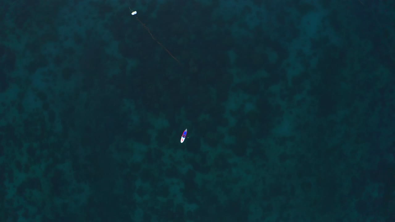 Top down aerial shot of a paddleboarder gliding over the calm, transparent waters of Koh Tao, Thailand, with coral reefs visible beneath the surface.