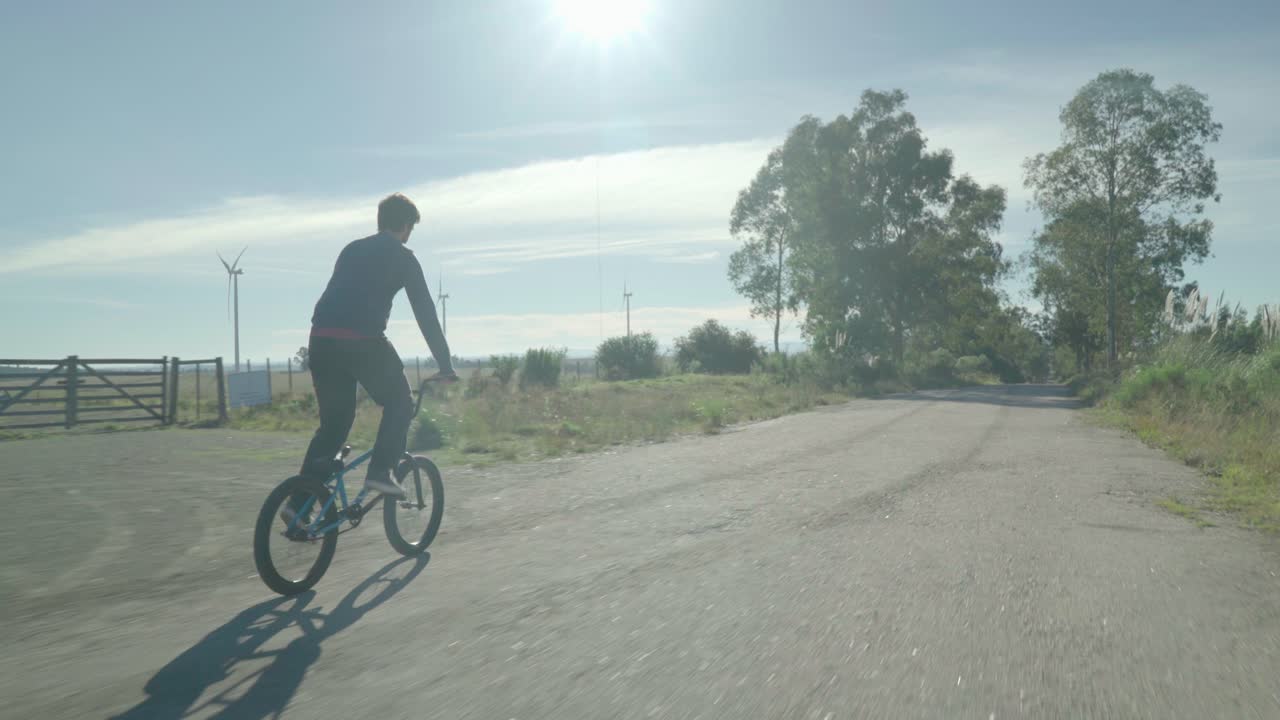 Young man rides on bmx down an abandoned road. On one side of the road you can see windmills producing wind power.