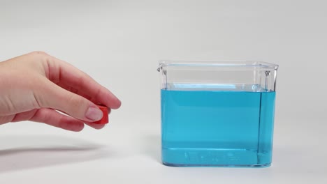 a hand drops a red object into a clear container with blue liquid, observing its sinking behavior