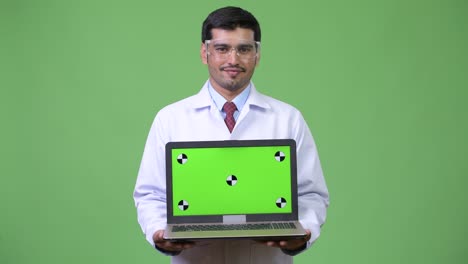 Young handsome Persian man doctor wearing protective glasses showing laptop