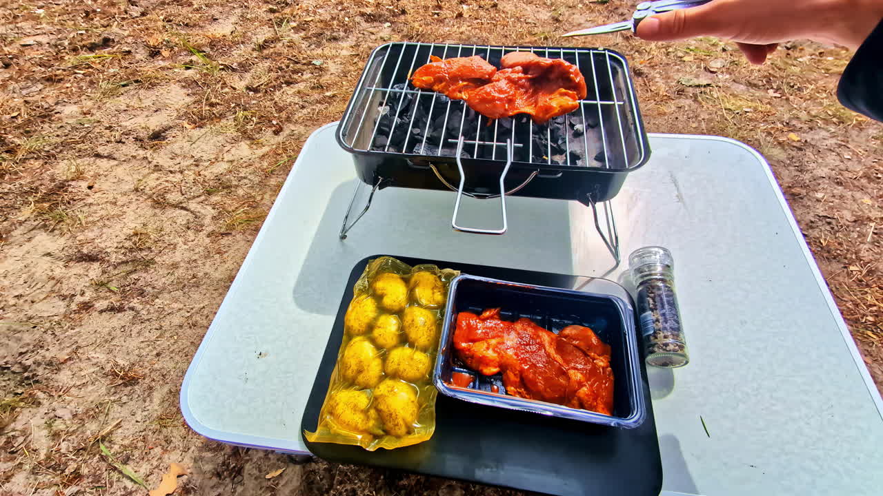POV shot shows a man preparing and grilling marinated chicken and potatoes on a small portable charcoal barbecue while camping outdoors