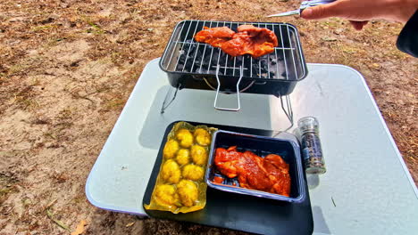 pov shot shows a man preparing and grilling marinated chicken and potatoes on a small portable charcoal barbecue while camping outdoors