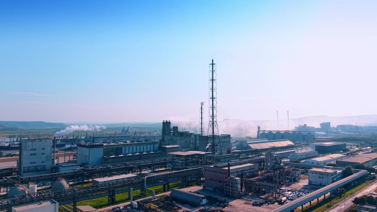 Moderne industrianlegg med et sentralt metallisk gittertårn. Lange stålrørledninger forbinder ulike bygninger på et prosessanlegg under en klar blå himmel.