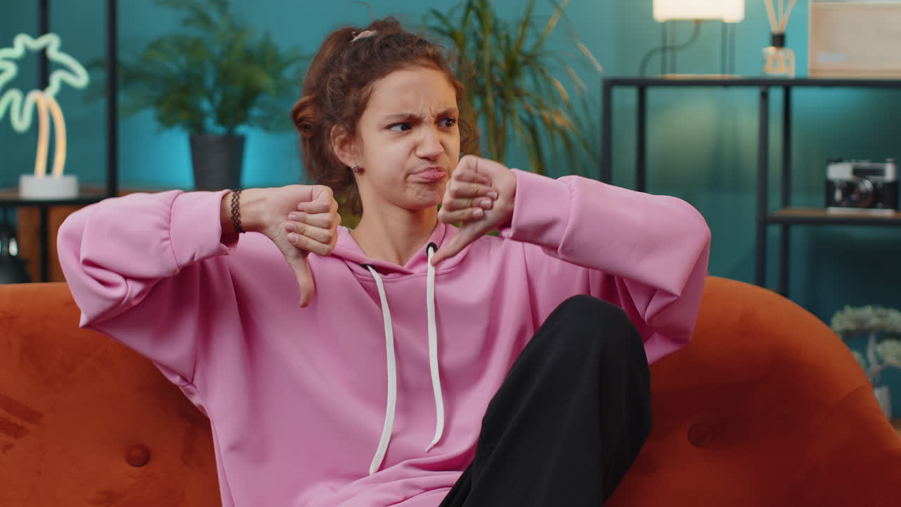 Upset school girl showing thumbs down dislike bad work disapproval dissatisfied feedback at home