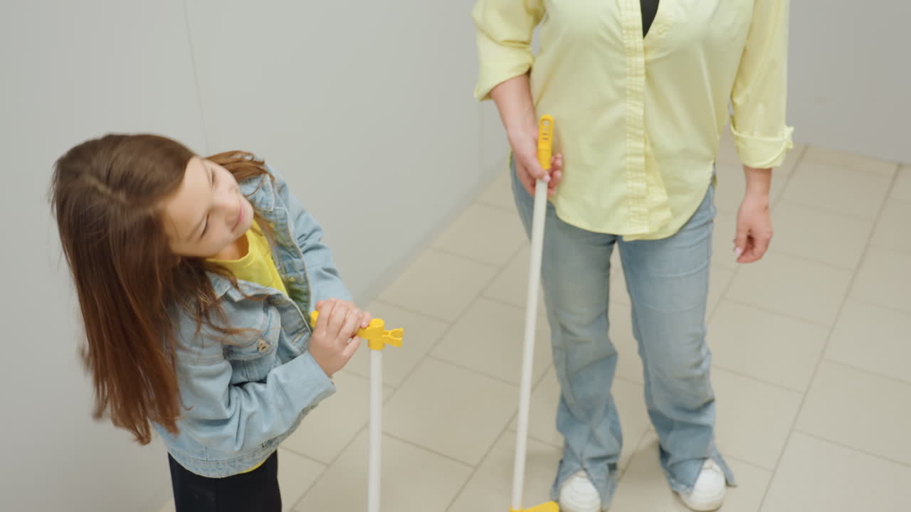 High angle view kid assisting mother in laundromat, excited holding yellow dustpan while parent sweeps tiled floor with broom, teamwork, chore moment, denim outfits, learning near stainless washers