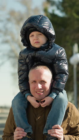 father carrying his child on his shoulders smiling, the father is wearing a brown jacket and beanie, and the child is in a black jacket with trees and light poles behind
