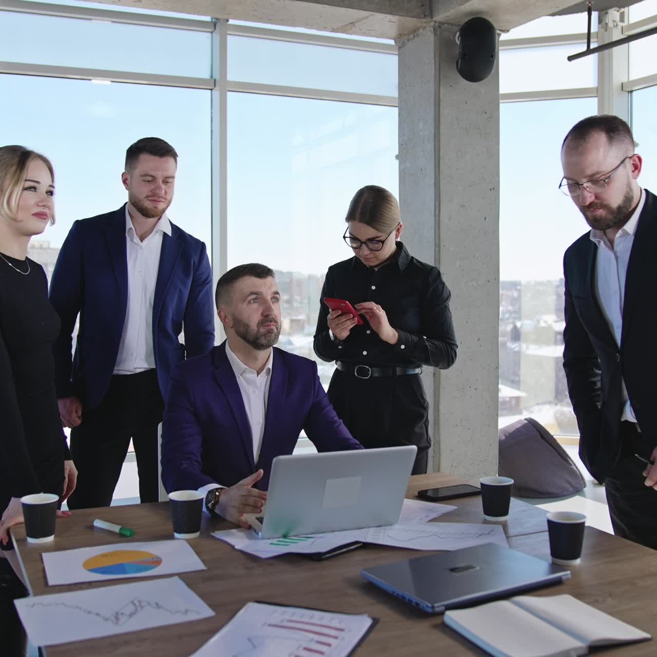 Business chief sits at the desk in front of laptop. Office team gathers around their leader. Panoramic windows with cityscape at backdrop