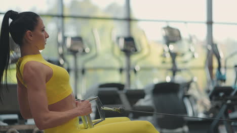 hispanic woman does exercises on the rowing machine. a girl sits behind a simulator is called a rowing machine. sitting pulls the weight of large windows and treadmills of the gym