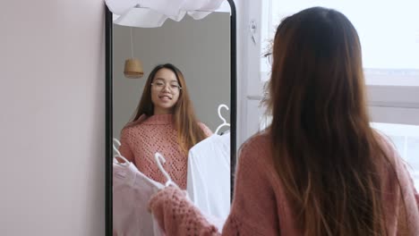 middle eastern young woman chooses clothes in front of a mirror in bedroom at house. girl think. hard choice routine of what clothes to wear. getting dressed