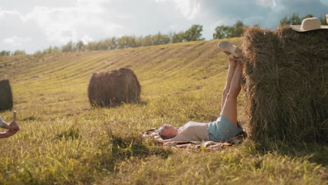 lady taking sister photos outdoors as she relaxes on mat, placing legs on hay in vast farmland, warm sunlight, countryside scenery, casual photography, and rustic setting with hat resting on hay bale