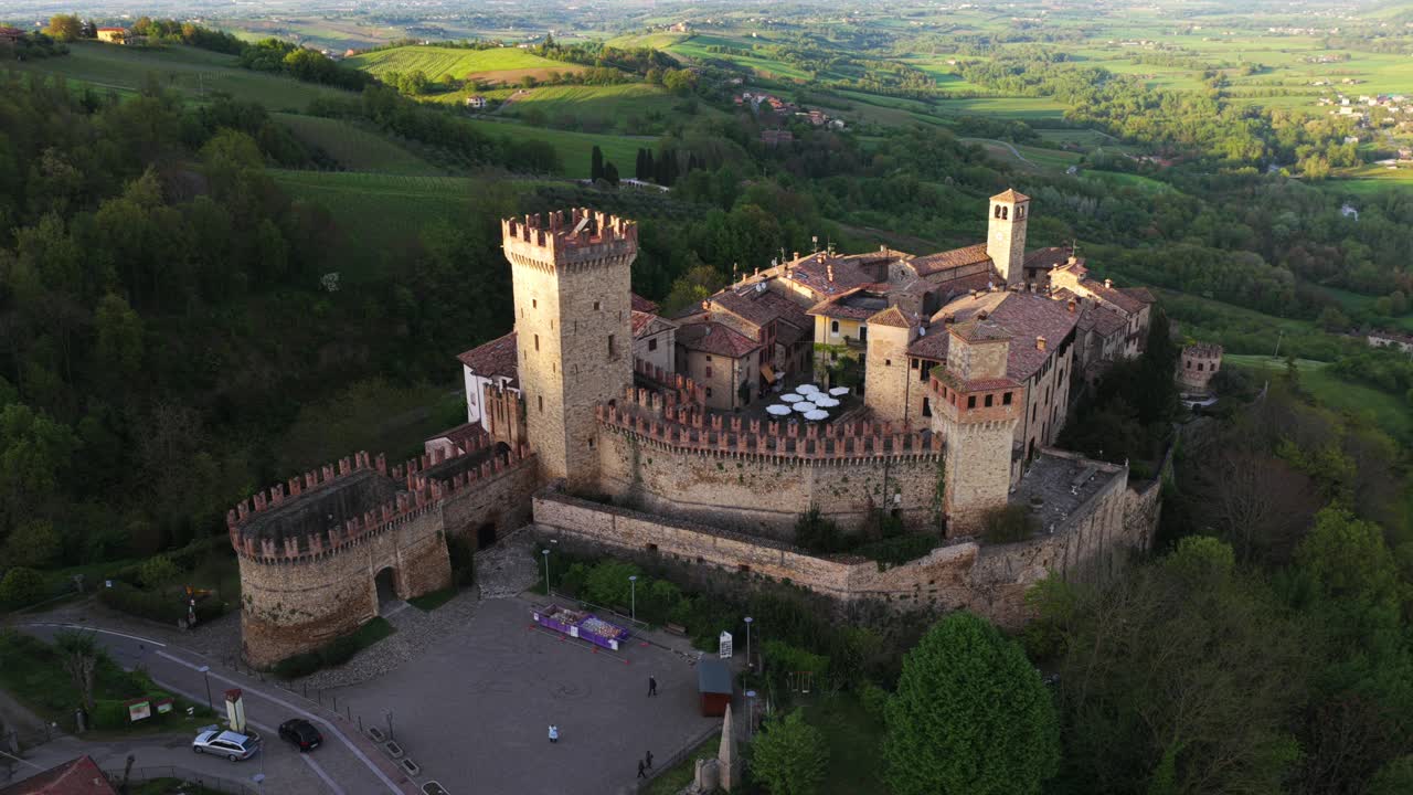 Aerial circling shot of Vigoleno historic castle at sunset, fortified medieval village on hilltop, surrounded by rolling green hills, Emilia-Romagna, Italy