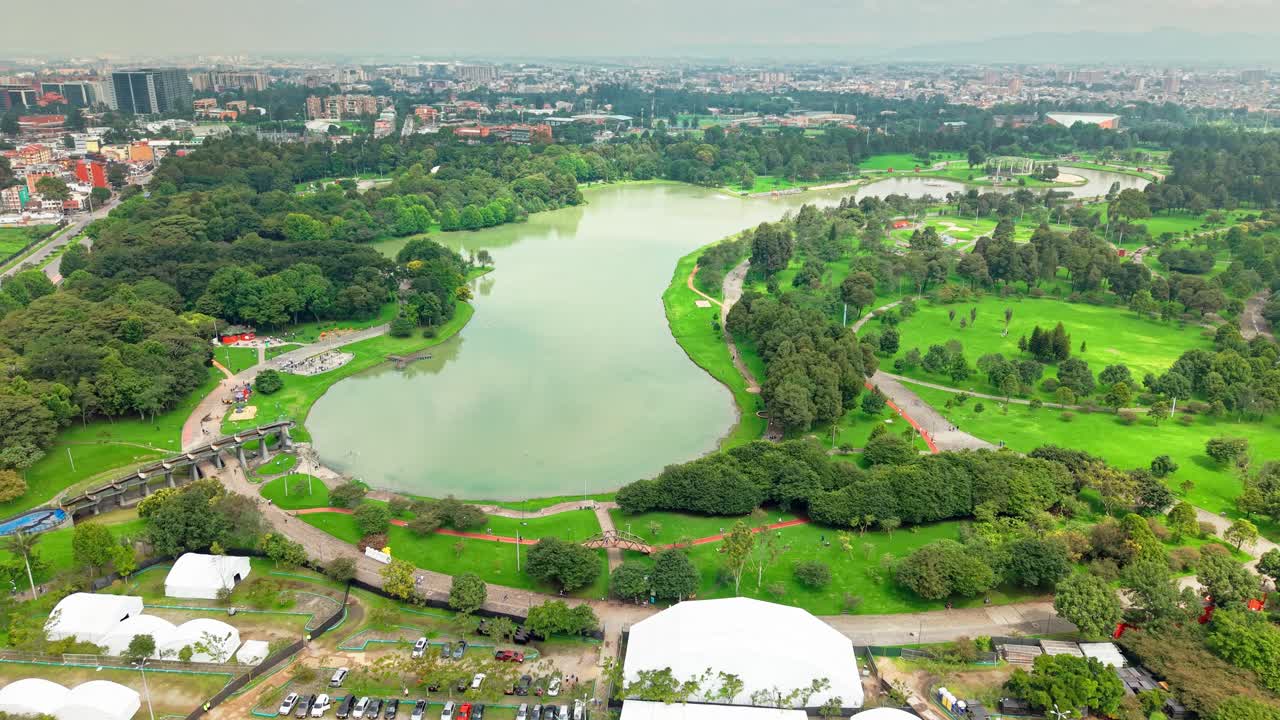 Receding aerial shot in Bogotá park showing green water, trees, and distant cityscape in high-angle view.