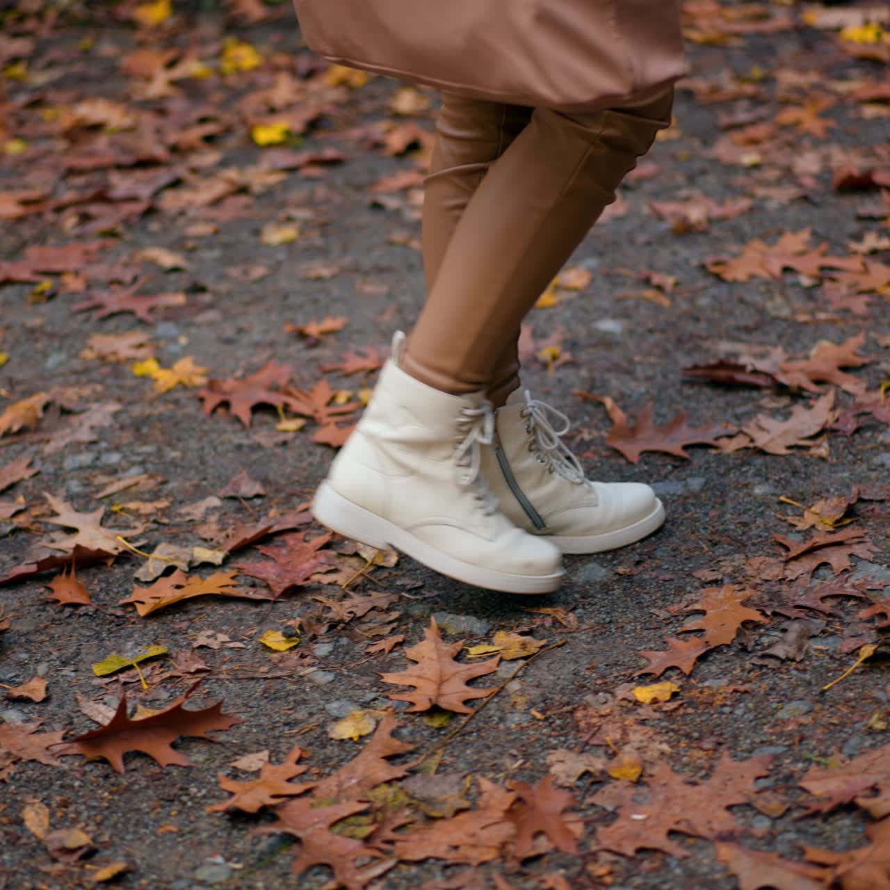 Unrecognized woman stepping by the road covered with leaves. Female legs in white boots having promenade in the autumn park