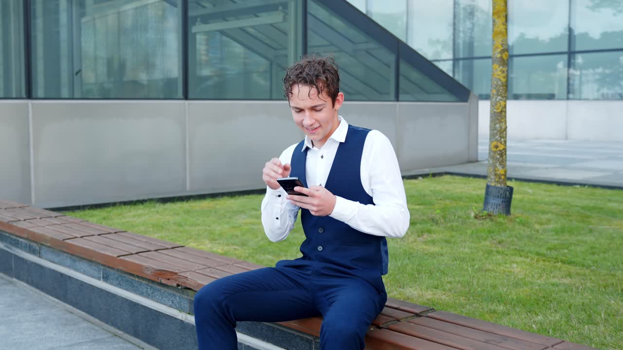 Young caucasian businessman sitting on urban bench, using mobile phone, browsing internet, managing business, embodying communication technology, and digital lifestyle outdoors, slow push in shot