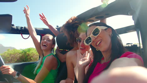 portrait of laughing female friends having fun posing for selfie in open top car on road trip