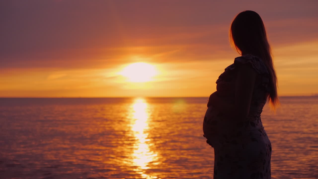 Giovane donna incinta con un pancione guarda il maestoso tramonto sul mare.