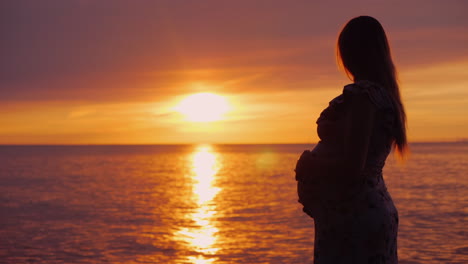 joven embarazada con una gran barriga mira la majestuosa puesta de sol sobre el mar